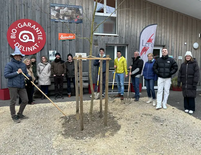Das Team pflanzt einen Baum vor dem Betriebsgebäude, an der Wand die Label von Delinat und Demeter